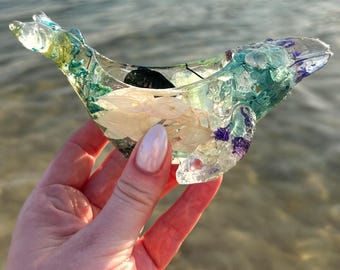 May include: A clear resin whale sculpture with embedded dried flowers and leaves. The whale is a light blue color with green and purple accents.
