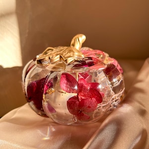 May include: A clear, pumpkin-shaped decorative container with a gold-coloured stem. The container is filled with dried red flowers and gold flakes. The container sits on a satin surface.