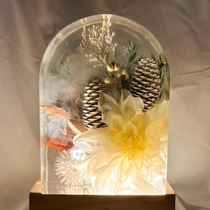 Puede incluir: Una pieza de arte decorativa de resina con una gran flor amarilla, piñas y follaje seco encerrados en resina transparente. La pieza está iluminada desde abajo y colocada sobre una base de madera.