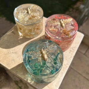 Op de afbeelding: Drie kleine, ronde, heldere hars potjes met goudkleurige bijen versieringen bovenop. De potjes zijn gevuld met gedroogde bloemen en bladgoud. Eén potje is geel, één is rood en één is turquoise. De potjes staan op een lichtgekleurd oppervlak.