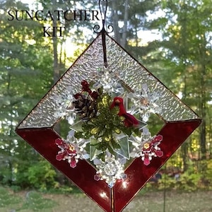 May include: A diamond-shaped suncatcher kit with a red border and clear, textured glass. The center features a red bird, pinecone, and decorative snowflakes. The text "Suncatcher Kit" is visible at the top.