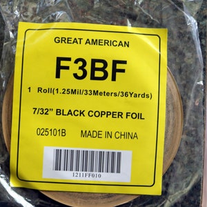 May include: A yellow label with black text reads "GREAT AMERICAN F3BF 1 Roll (1.25Mil/33Meters/36Yards) 7/32" BLACK COPPER FOIL 025101B MADE IN CHINA 1211FF010". A barcode is below the text. The label is attached to a roll of copper foil.