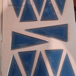 May include: A collection of blue triangular glass tiles. Each tile has a beveled edge, creating a 3D effect. The tiles are arranged on a white background, suggesting a craft or mosaic project. The image includes text in the upper left corner.