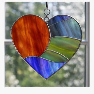 May include: A heart-shaped stained glass suncatcher titled Precut Heart Scene Kit # 1. The piece features a large orange section, a blue bottom section, and a multi-colored right side with blue and green segments, all joined by silver metal solder lines.