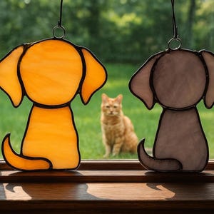 May include: Two stained glass dog suncatchers, one amber and one gray, hang in front of a window. A ginger cat sits in the background on the green grass. The dog silhouettes have large ears and curled tails.