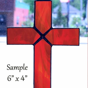 May include: A stained glass cross in shades of red and orange, with a dark blue border. The cross is approximately 6 inches tall and 4 inches wide. It has a decorative metal loop for hanging. The word "Sample" is written on the cross.
