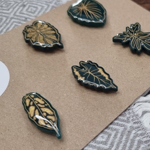 May include: Five green resin leaf-shaped magnets with gold leaf detailing. The magnets are on a brown surface.