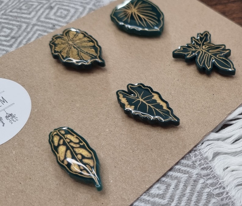 May include: Five green resin leaf-shaped magnets with gold leaf detailing. The magnets are on a brown surface.