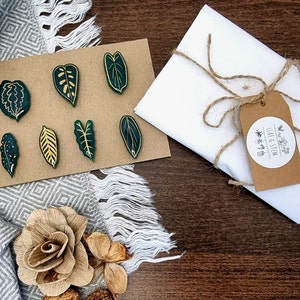 May include: A set of seven green leaf-shaped pins with gold details, packaged in a white envelope with a brown paper tag that reads "Leaf & Stem".