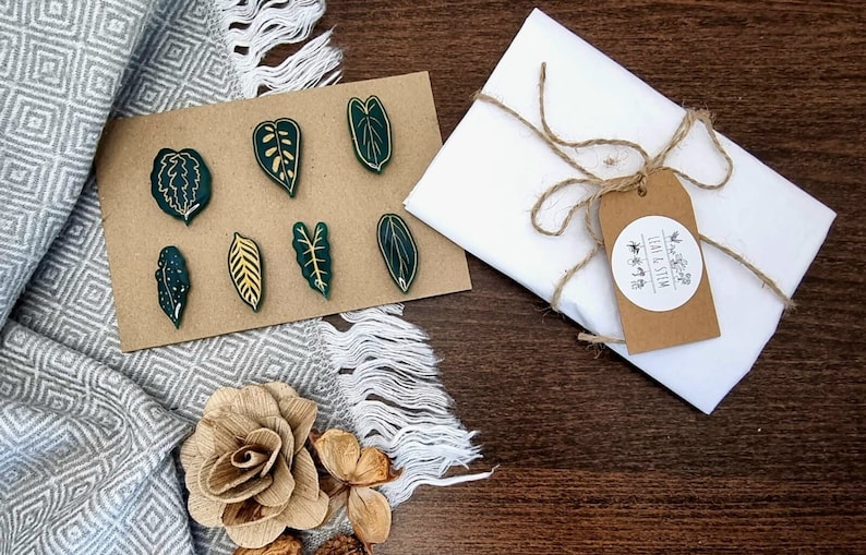 May include: A set of seven green leaf-shaped pins with gold details, packaged in a white envelope with a brown paper tag that reads "Leaf & Stem".