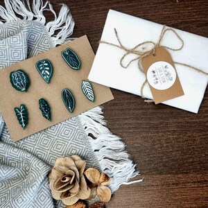 May include: A set of seven green leaf-shaped pins on a brown background. The pins are arranged in a semi-circle. The background is a brown fabric with a white and gray pattern. The pins are packaged in a white envelope with a brown paper tag that says "Leaf & Stem".