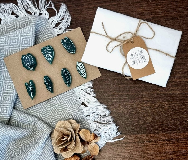 May include: A set of seven green leaf-shaped pins on a brown background. The pins are arranged in a semi-circle. The background is a brown fabric with a white and gray pattern. The pins are packaged in a white envelope with a brown paper tag that says "Leaf & Stem".