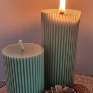 May include: Two green pillar candles with a ribbed design. The larger candle is lit and the smaller candle is unlit. The candles are on a wooden surface with a decorative flower and a nut.