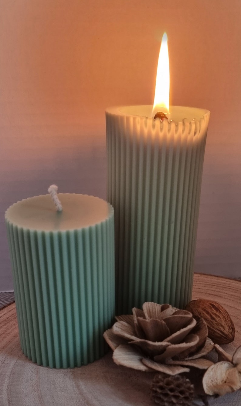 May include: Two green pillar candles with a ribbed design. The larger candle is lit and the smaller candle is unlit. The candles are on a wooden surface with a decorative flower and a nut.