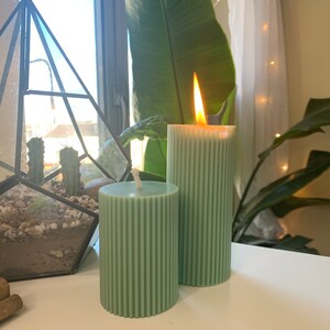 May include: Two green pillar candles with a ribbed texture. One candle is lit and has a flame.