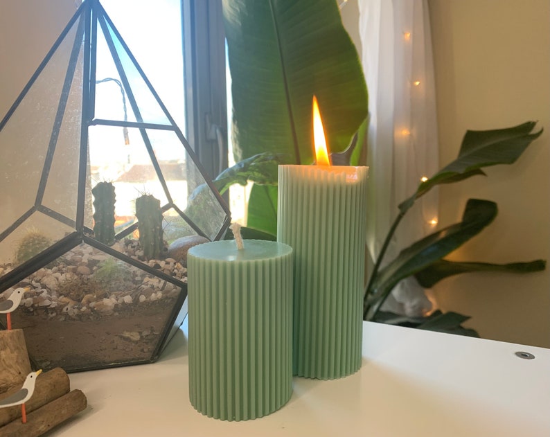 May include: Two green pillar candles with a ribbed texture. One candle is lit and has a flame.