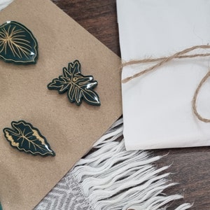May include: Set of five green and gold leaf-shaped pins. The pins are arranged on a brown card with the text "Leaf & Stem" on a brown tag.
