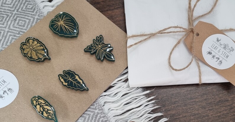 May include: Set of five green and gold leaf-shaped pins. The pins are arranged on a brown card with the text "Leaf & Stem" on a brown tag.