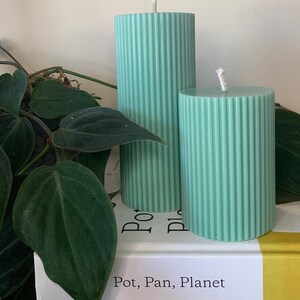 May include: Two green pillar candles with a ribbed texture. The candles are on top of two books. The top book has the text "Pot, Pan, Planet" on the cover. The bottom book has the text "FITWAFFLE'S Baking It Easy" on the cover.