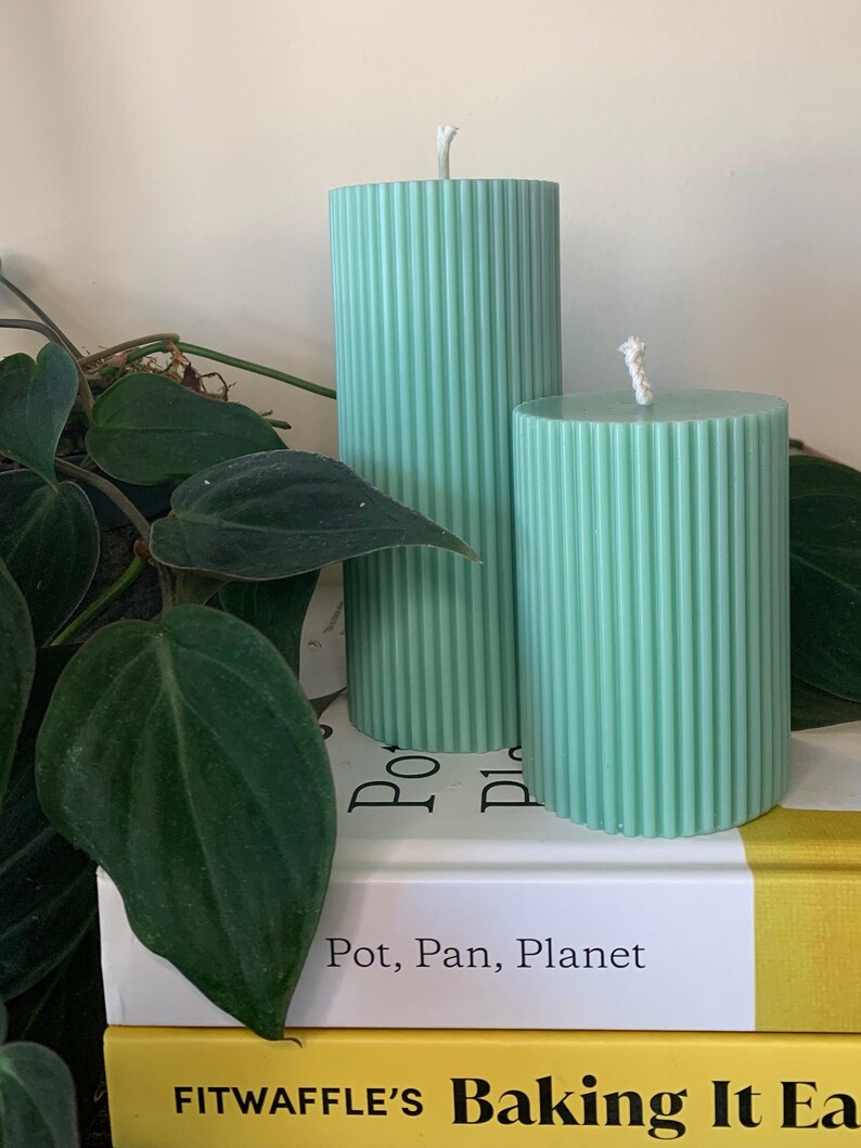 May include: Two green pillar candles with a ribbed texture. The candles are on top of two books. The top book has the text "Pot, Pan, Planet" on the cover. The bottom book has the text "FITWAFFLE'S Baking It Easy" on the cover.