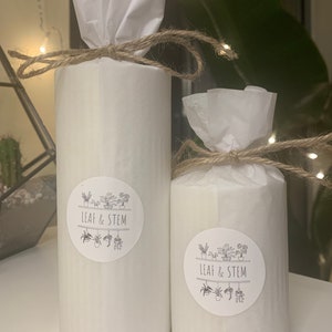 May include: Two white cylindrical candles wrapped in white tissue paper and tied with twine. The candles have black and white stickers with the text "LEAF & STEM" and a graphic of potted plants.