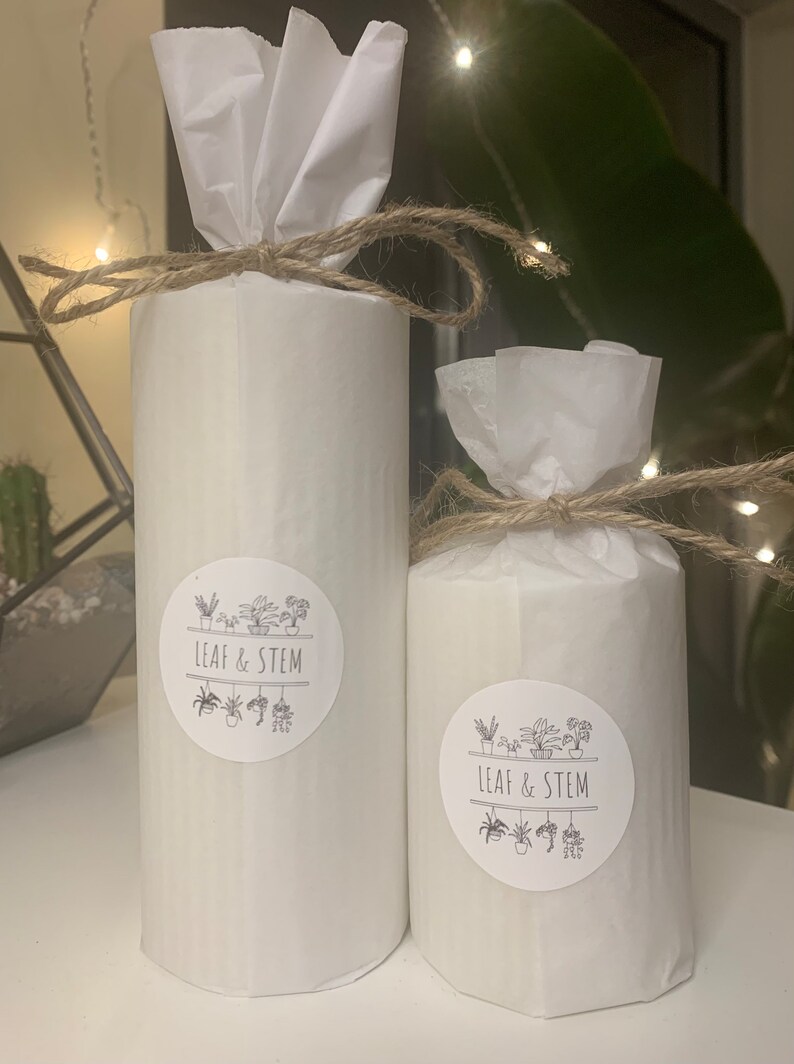 May include: Two white cylindrical candles wrapped in white tissue paper and tied with twine. The candles have black and white stickers with the text "LEAF & STEM" and a graphic of potted plants.