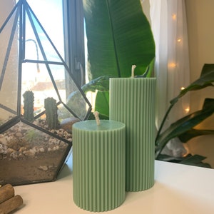 May include: Two green pillar candles with a ribbed texture. The candles are on a white surface.