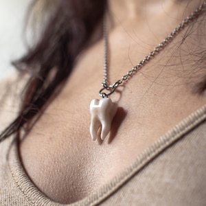 May include: A pendant necklace featuring a detailed, off-white tooth charm. The tooth is suspended from a silver-colored chain. The necklace is worn against a neutral-toned top, creating a contrast that highlights the pendant.