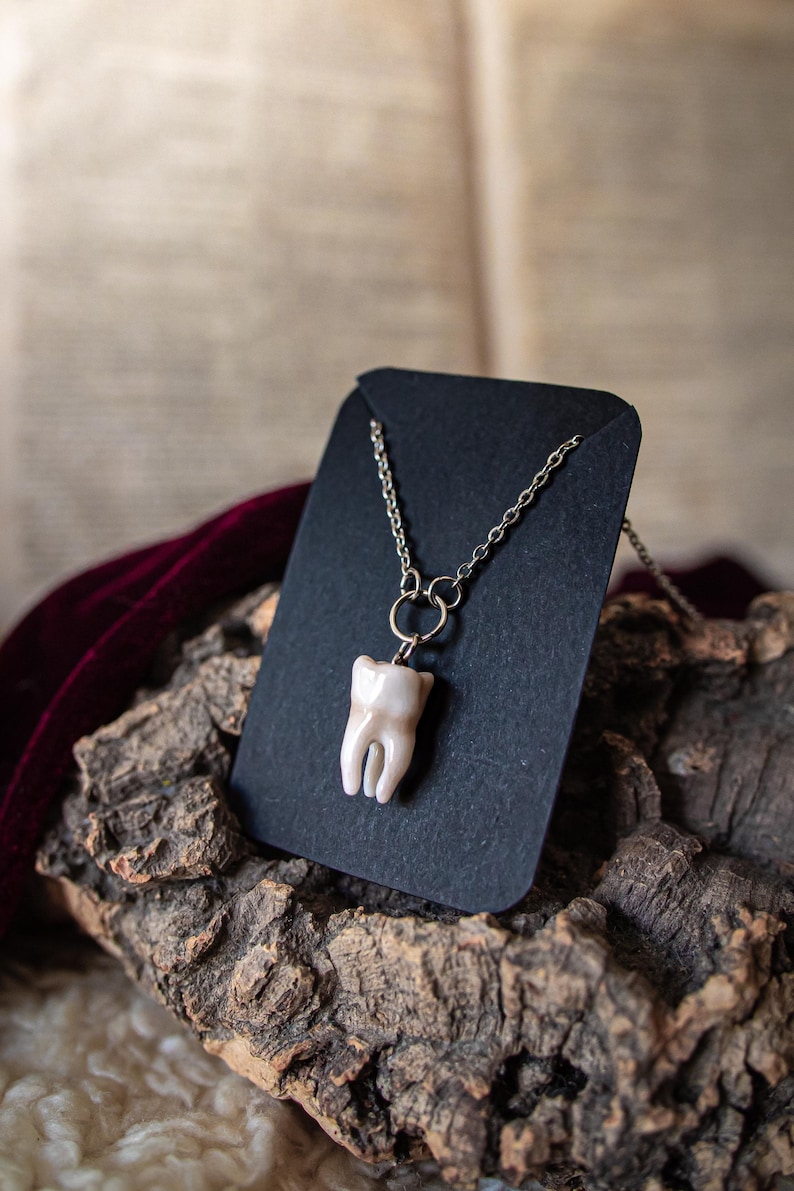May include: A tooth-shaped pendant necklace. The pendant is a light beige color, with a silver-colored chain. The necklace is displayed on a black card, resting on a piece of wood. The background is blurred.