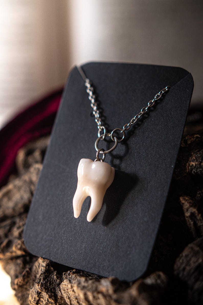 May include: A pendant necklace featuring a realistic tooth charm. The tooth is off-white and detailed, suspended from a silver-colored chain. The necklace is displayed on a black card against a textured, dark background.