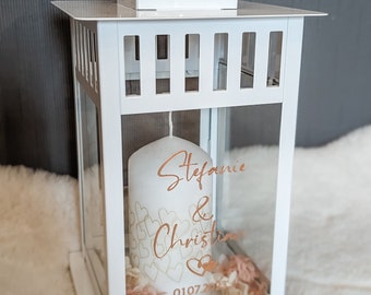 May include: White metal lantern with a glass panel, containing a white candle with the names "Stefanie & Christina" and the date "010720" written in gold lettering. The candle is surrounded by small pink flowers.