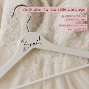 Könnte beinhalten: Ein weißer Holzkleiderbügel mit dem Wort "Braut" in schwarzer Schrift. Der Kleiderbügel hängt an einem weißen Brautkleid mit floralen Spitzendetails. Der Text auf dem Bild lautet "Aufkleber für den Kleiderbügel" und "IN VERSCHIEDENEN SCHRIFTFARBEN VERSCHIEDENE TEXTE ZUR AUSWAHL."