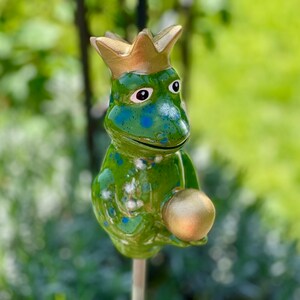 Könnte beinhalten: Eine grüne Froschfigur mit blauen Flecken, die eine goldene Krone trägt und eine goldene Kugel hält. Der Frosch sitzt auf einem Stock.