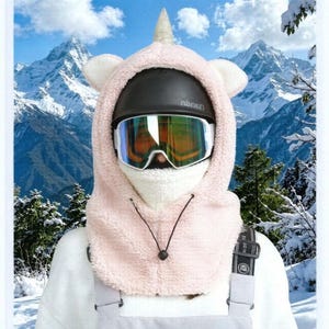 May include: A light pink fleece balaclava featuring a unicorn horn and ears, worn over a black helmet and reflective ski goggles. The hood includes a white textured neck warmer section and a black drawstring cord with a toggle for adjustment.