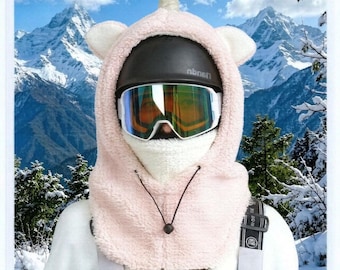 Könnte beinhalten: Eine hellrosa Fleece-Sturmhaube mit Einhornhorn und Ohren, getragen über einem schwarzen Helm und reflektierender Skibrille. Die Kapuze verfügt über einen weißen, strukturierten Nackenwärmer und eine schwarze Kordel mit einem Stopper.