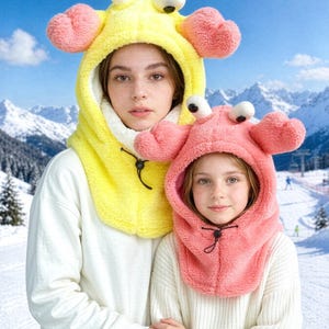 K&ouml;nnte beinhalten: Eine leuchtend gelbe Pl&uuml;schkapuze mit krabbenartigen Augen und rosa Scheren, ausgestattet mit wei&szlig;em Innenfutter und einem schwarzen Kordelzug. Daneben eine passende rosa Pl&uuml;schkapuze mit wei&szlig;en Augen und einem schwarzen Kordelzug.