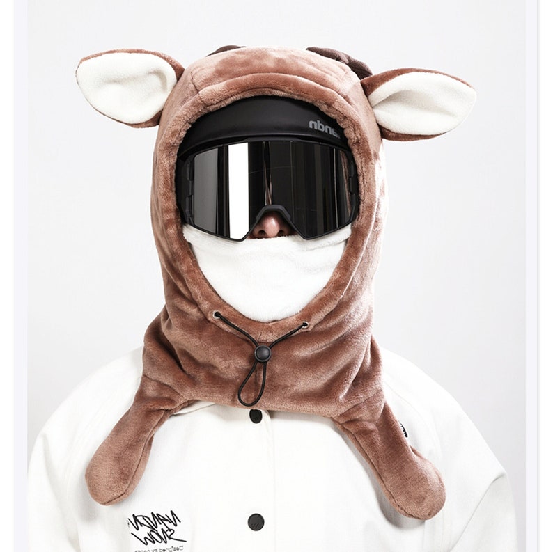 May include: A brown plush animal-themed hood featuring large ears, worn over a white face mask and black mirrored ski goggles. The hood includes a black drawstring with a toggle closure at the neck, providing full head and neck coverage.