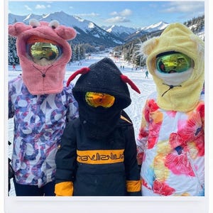 K&ouml;nnte beinhalten: Drei Personen tragen Fleece-Sturmhauben mit tierischen Kapuzen und Skibrillen. Die linke Kapuze ist rosa mit krebsartigen Augen, die mittlere ist schwarz mit roten Ohren und die rechte ist gelb mit b&auml;renartigen Ohren. Sie tragen gemusterte Skijacken.