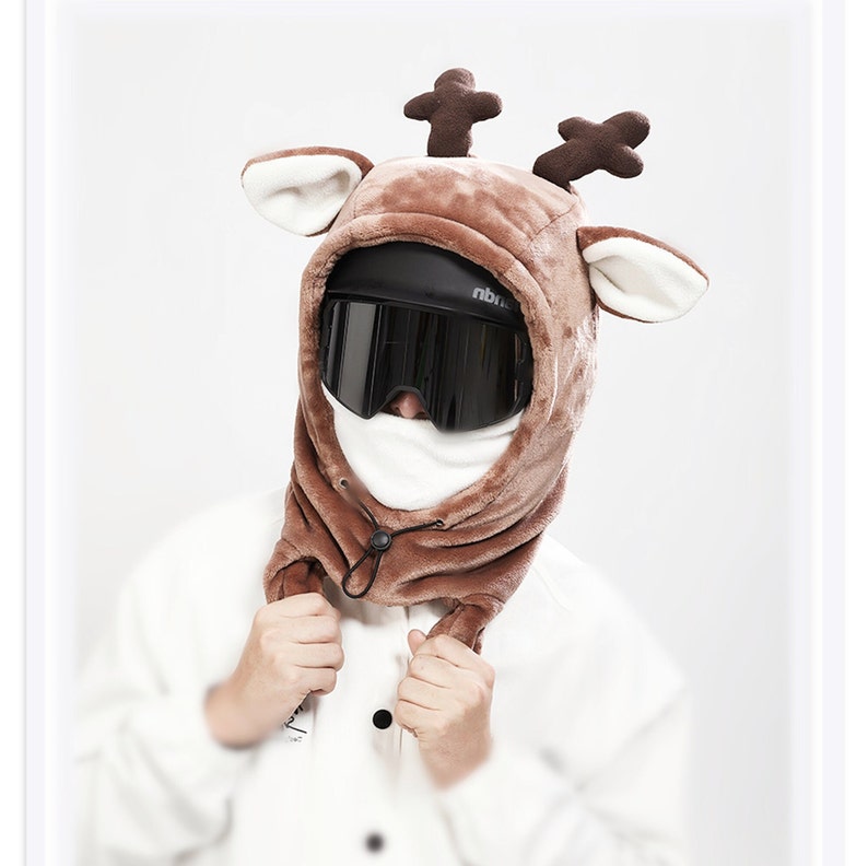 May include: A brown plush reindeer hood featuring dark brown antlers and cream-colored inner ears. The hood is worn over a black helmet with dark tinted ski goggles and a white face covering, secured with a black drawstring toggle at the neck.