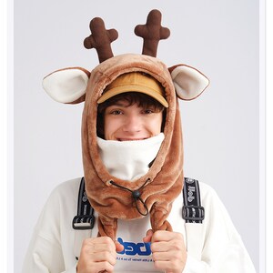 May include: A brown plush reindeer hood featuring dark brown antlers and white-lined ears. The hood includes a white fleece neck warmer section with a black drawstring toggle closure, worn over a tan baseball cap and a white sweatshirt.