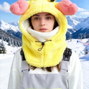 K&ouml;nnte beinhalten: Eine leuchtend gelbe Pl&uuml;sch-Fleece-Sturmhaube mit zwei gro&szlig;en wei&szlig;en Cartoon-Augen auf der Oberseite und zwei rosafarbenen, abgerundeten seitlichen Anh&auml;ngseln. Die Kapuze verf&uuml;gt &uuml;ber ein wei&szlig;es Innenfutter und einen schwarzen Kordelzug.