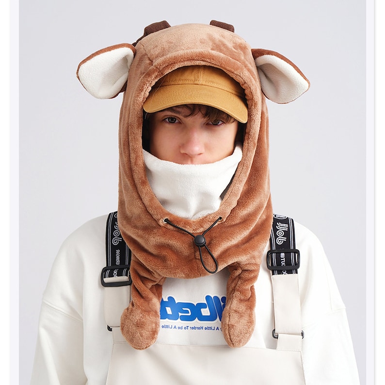 May include: A brown plush animal-themed hood featuring soft ears and small horns on top. The hood includes a white fleece-lined interior, a drawstring closure with a black toggle, and two long side flaps that hang down. The fabric is textured.