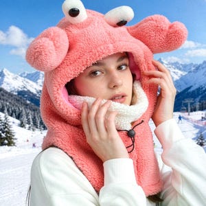 K&ouml;nnte beinhalten: Eine korallenrosa Pl&uuml;schkapuze mit zwei gro&szlig;en, cartoonartigen Augen auf der Oberseite und zwei seitlichen Ausw&uuml;chsen, die an Krabbenscheren erinnern. Das Innere ist mit wei&szlig;em Fleece gef&uuml;ttert und verf&uuml;gt &uuml;ber einen schwarzen Kordelzug mit Stopper.