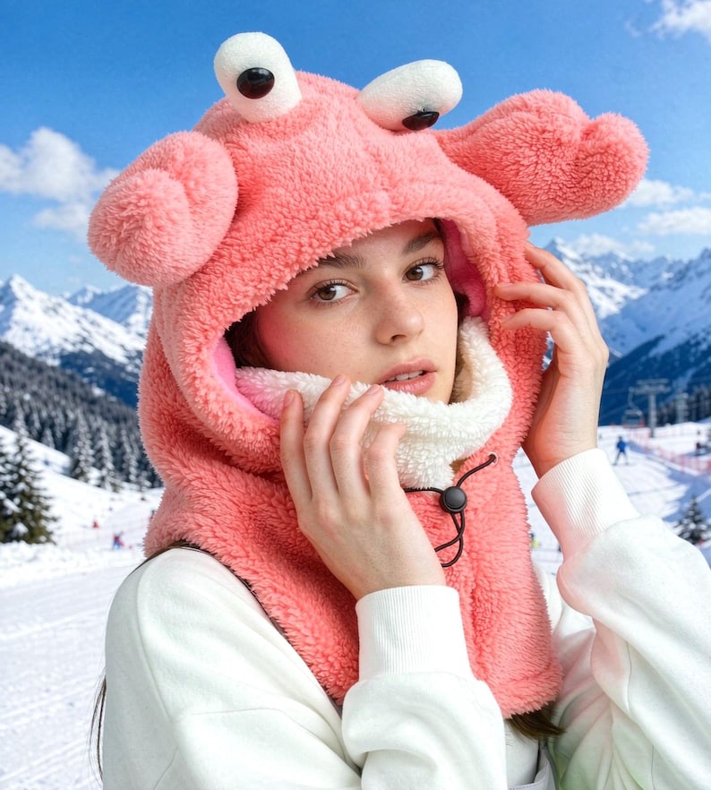 K&ouml;nnte beinhalten: Eine korallenrosa Pl&uuml;schkapuze mit zwei gro&szlig;en, cartoonartigen Augen auf der Oberseite und zwei seitlichen Ausw&uuml;chsen, die an Krabbenscheren erinnern. Das Innere ist mit wei&szlig;em Fleece gef&uuml;ttert und verf&uuml;gt &uuml;ber einen schwarzen Kordelzug mit Stopper.