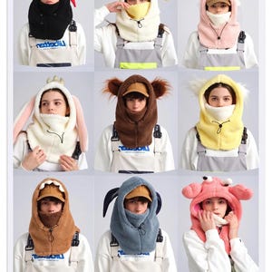 K&ouml;nnte beinhalten: Eine Kollektion von neun Balaclava-Kapuzen aus Pl&uuml;schfleece mit verschiedenen tierinspirierten Designs, darunter ein schwarzer Teufel, eine gelbe Ente, ein rosa Einhorn, ein wei&szlig;er Hase, ein brauner B&auml;r, ein gelbes K&uuml;ken, ein braunes Monster, ein blauer Hund und eine rosa Krabbe.