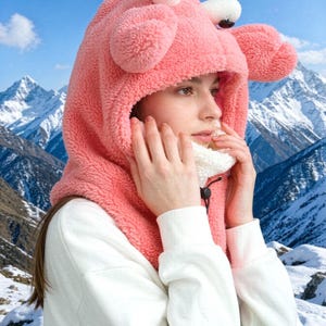 K&ouml;nnte beinhalten: Ein rosafarbener Pl&uuml;sch-Kapuzenschal mit zwei hervorstehenden Augen und zwei abgerundeten Forts&auml;tzen an der Oberseite. Die Kapuze besteht aus flauschigem, strukturiertem Material und hat ein wei&szlig;es Innenfutter sowie einen schwarzen Kordelzug am Hals.