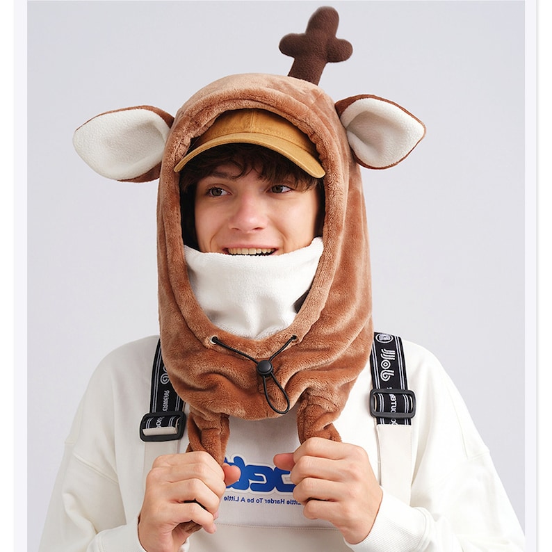 May include: A brown plush hood featuring deer-like ears and a single antler on top. The hood includes a white fleece neck warmer, a black drawstring toggle for adjustment, and is worn over a tan baseball cap and a white long-sleeve shirt.