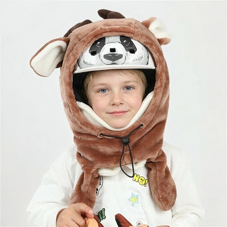 May include: A brown plush animal-themed helmet cover featuring large ears and antlers, designed to fit over a white helmet with a black and white panda face graphic on the front. The cover includes a black drawstring toggle for a secure fit.