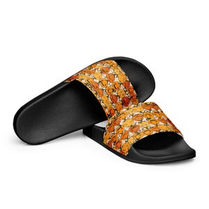 May include: Black slide sandals with a repeating pattern of orange and yellow cartoon faces. The sandals have a black sole and a wide strap across the top of the foot. The design is playful and colorful.