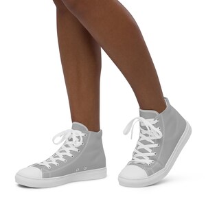May include: A pair of high-top sneakers in a solid light gray color. The sneakers have white laces, a white rubber toe cap, and a white sole. The shoes are shown from a low angle.